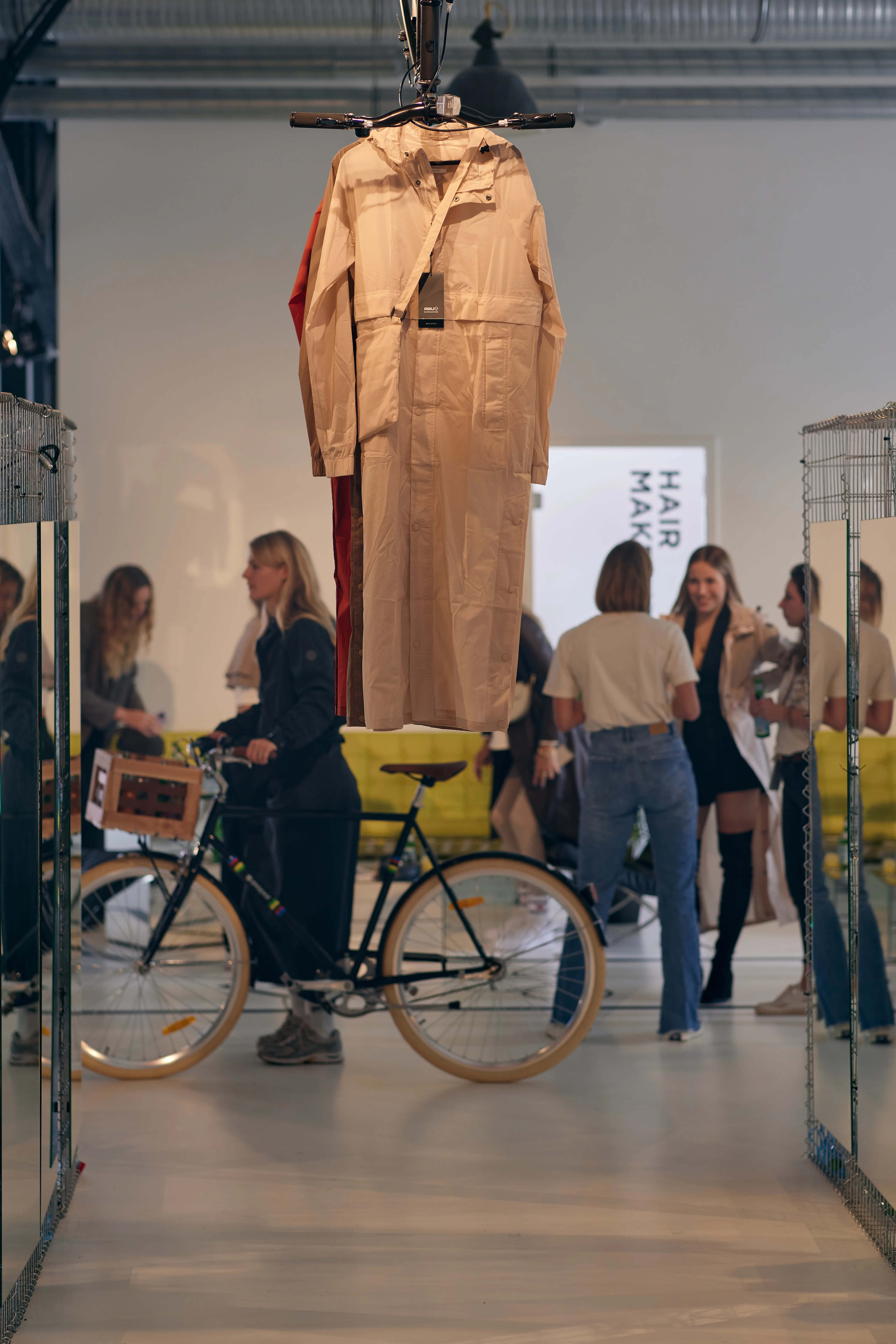 Beige AGU rain coat hanging from the ceiling above a bicycle, with visitors mingling in the bright showroom behind.
