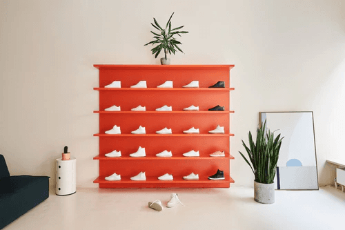 A vibrant red wall-mounted shoe rack displaying white sneakers in a stylish room with minimalistic decor, featuring a potted plant on top, a large mirror, and sleek greenery on the floor.