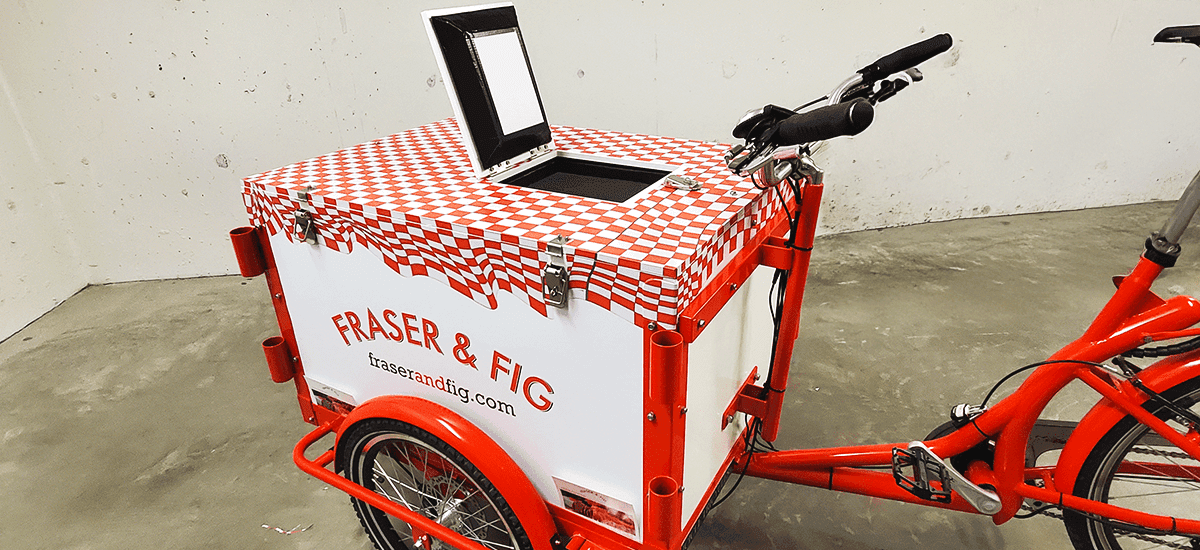 a red-and-white bicycle with fridge box attachment and vinyl wrap on top, reading Fig & Fraser on the sides and front