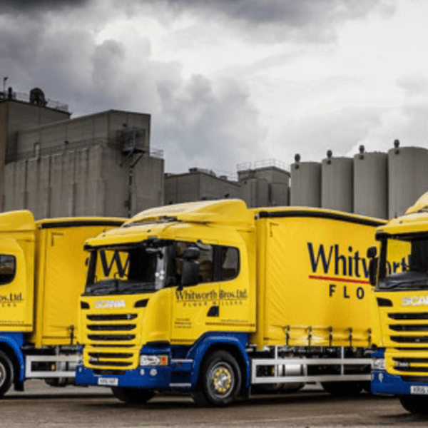 Three yellow delivery trucks parked in front of a warehouse under a cloudy sky.