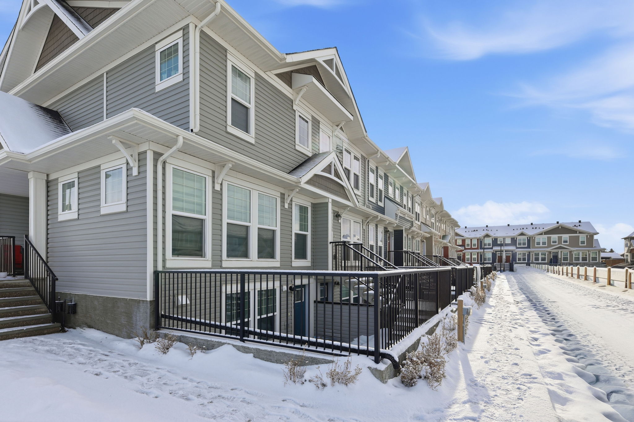 Row of modern two-story townhomes with grey siding, white trim, black metal patio railings, and a snow-dusted walkway under a blue sky in Falconridge, NE Calgary