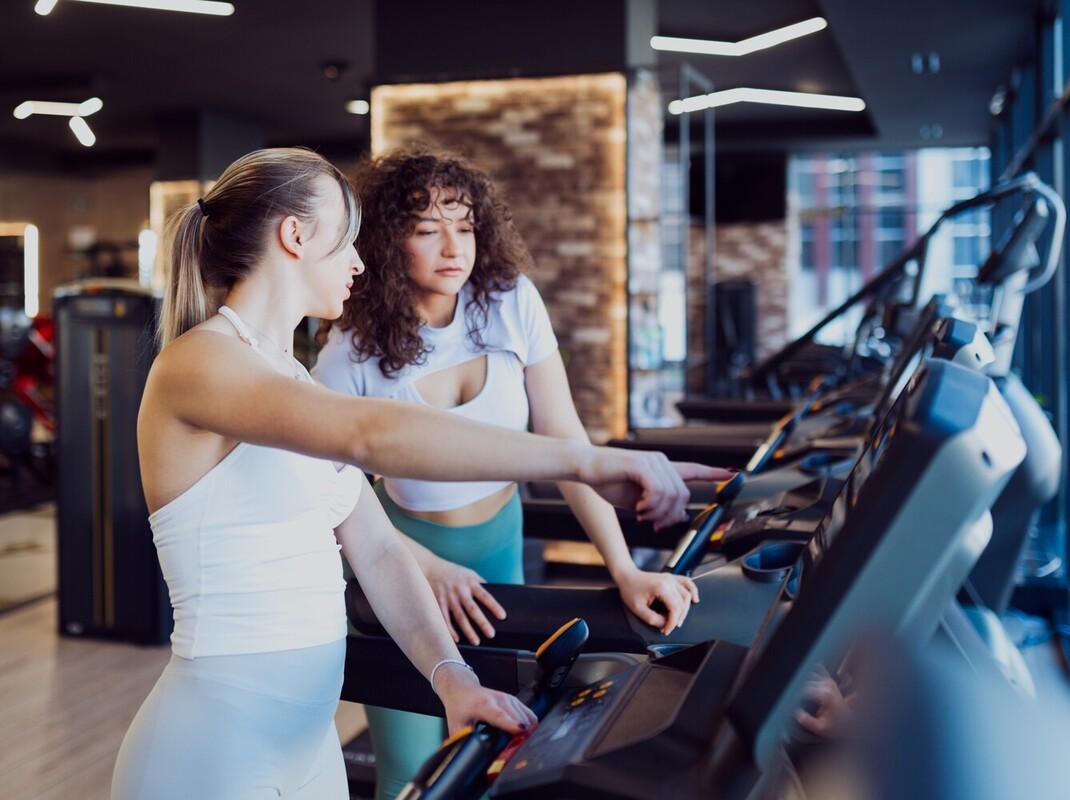 woman helping her friend select speed settings for a HIIT treadmill workout for weight loss at the gym