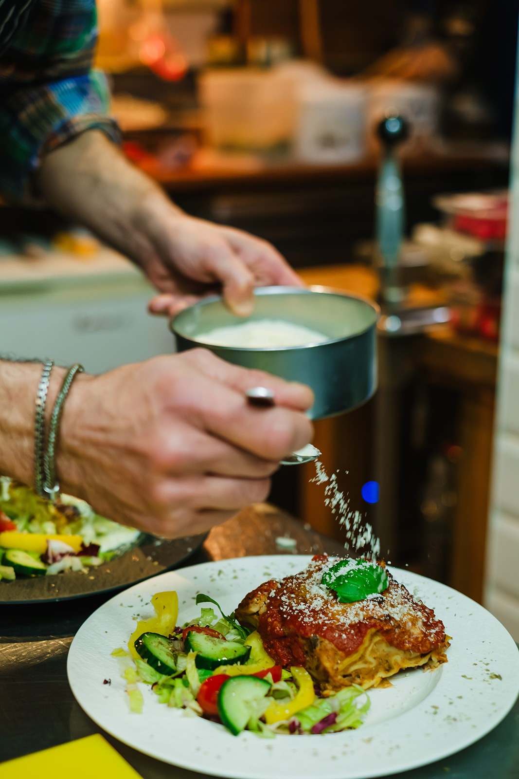 A chef wearing a striped apron and plaid shirt carefully plates fresh ravioli, using a spoon to arrange pieces from a pan in a warmly lit kitchen setting.
