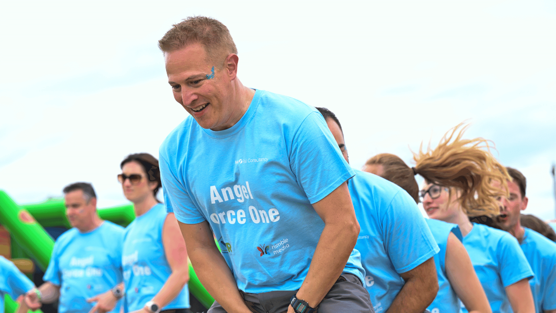 Outdoor team-building event with participants wearing matching blue shirts, taking part in inflatable obstacle courses and games, smiling and cheering, with the words 'ENTER A TEAM' in large bold white and green text across the image.