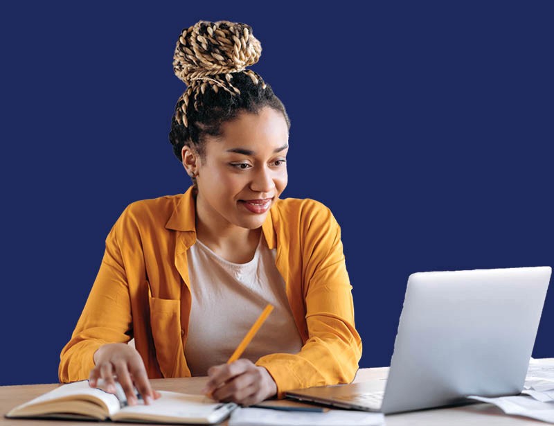 Young woman studies at a desk, writing in a notebook while looking at a laptop against a solid blue background.