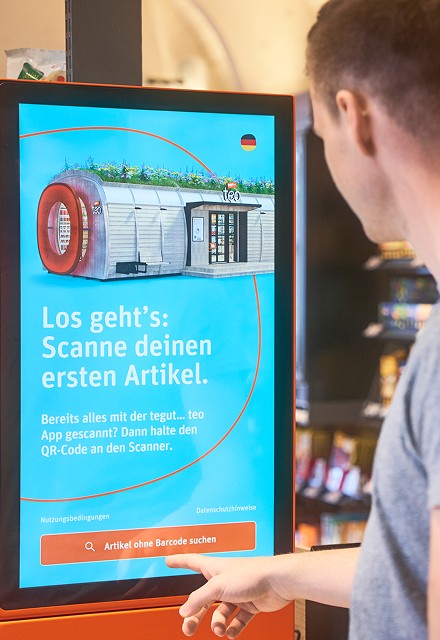 Innenansicht des tegut… teo mit einer großen Selbstbedienungskasse im Vordergrund und dem gewölbten, hölzernen Innenraum mit gut gefüllten Regalen im Hintergrund.