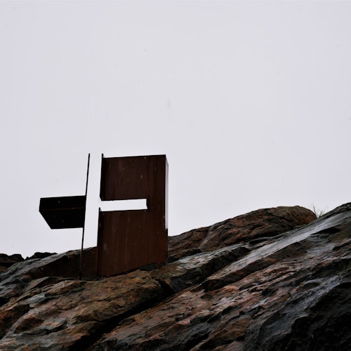 Metallinen abstrakti veistos kivisellä mäellä pilvisen taivaan alla.