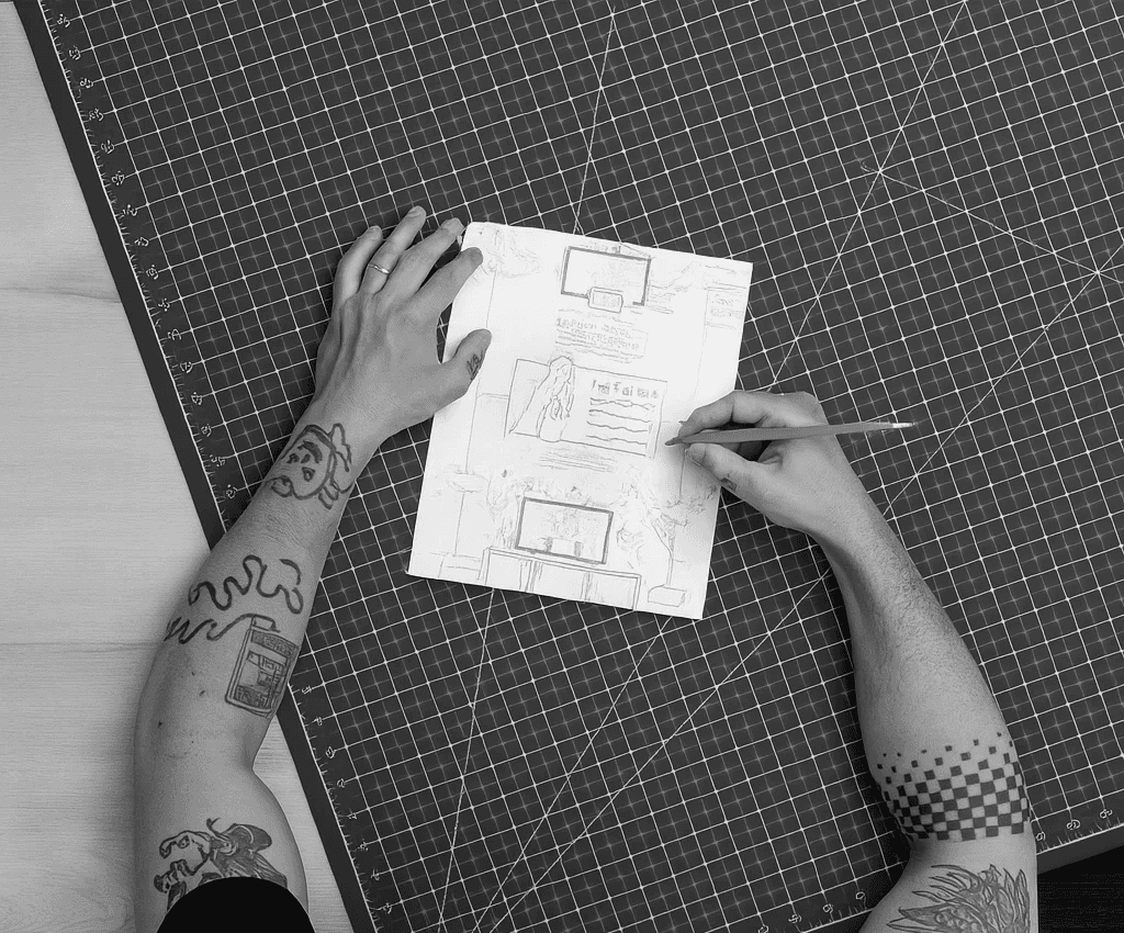 A bird's eye view of two tattooed arms holding and drawing on a piece of paper with a grid cutting mat underneath.