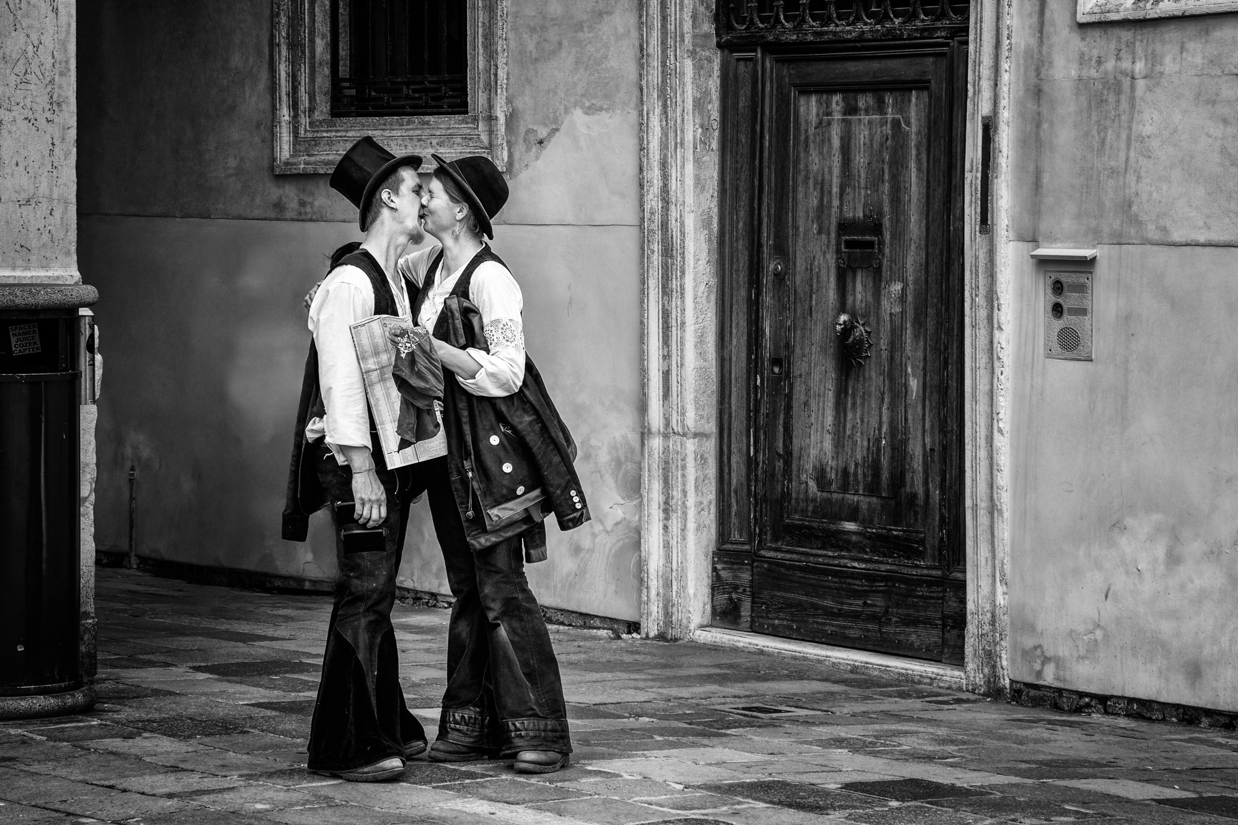 Two people dressed in vintage costumes kissing on a narrow street with a worn wooden door in the background