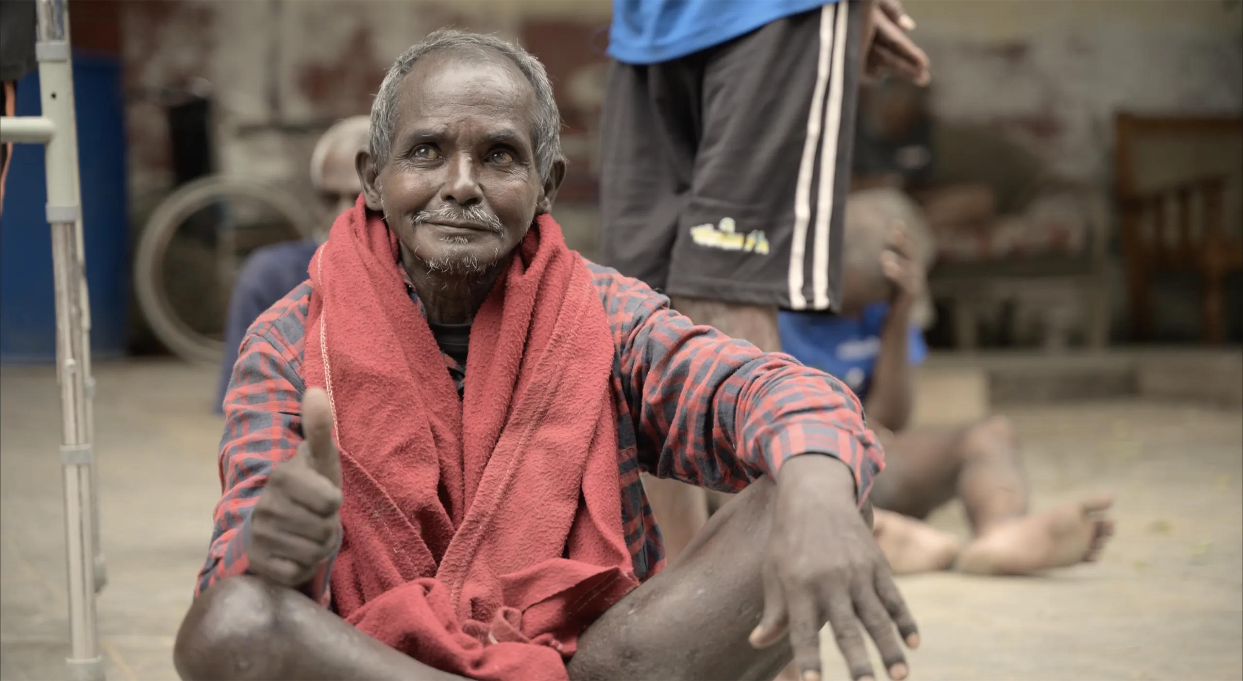 A warm and hopeful portrait of an elderly man with a red scarf giving a thumbs up in a communal setting in Bangalore India.