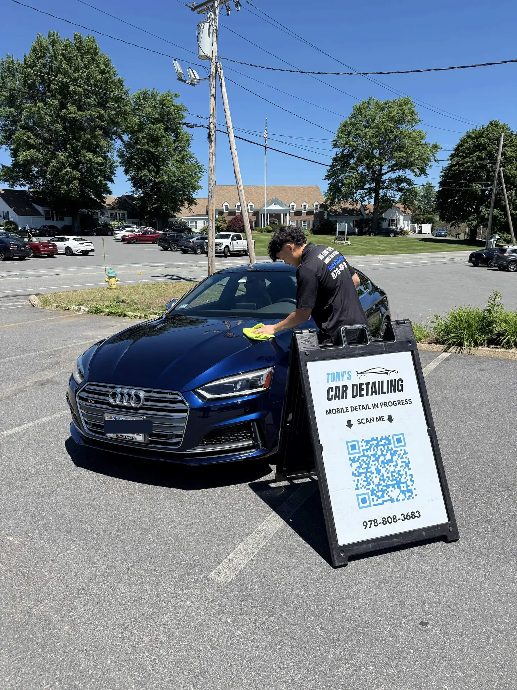A mobile detailer wiping down the hood of a shiny, dark blue Audi sedan in a parking lot on a sunny day next to a "Tony's Car Detailing" A-frame sign with a QR code.