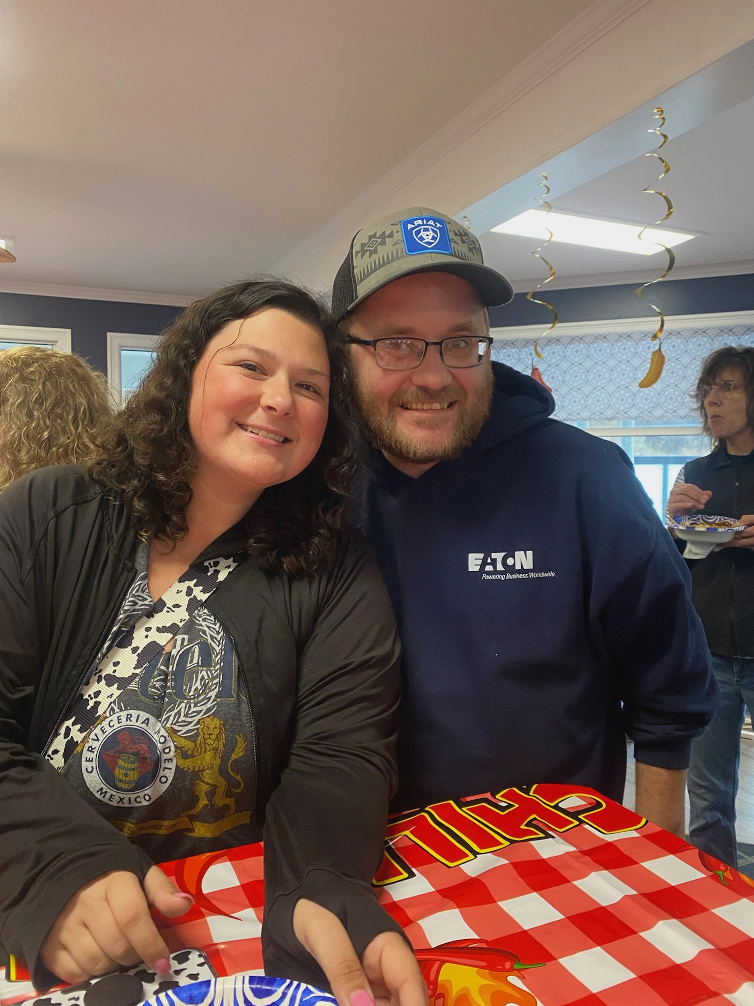A smiling couple poses at a party beside a checkered tablecloth, with festive decorations hanging in the background and guests mingling.