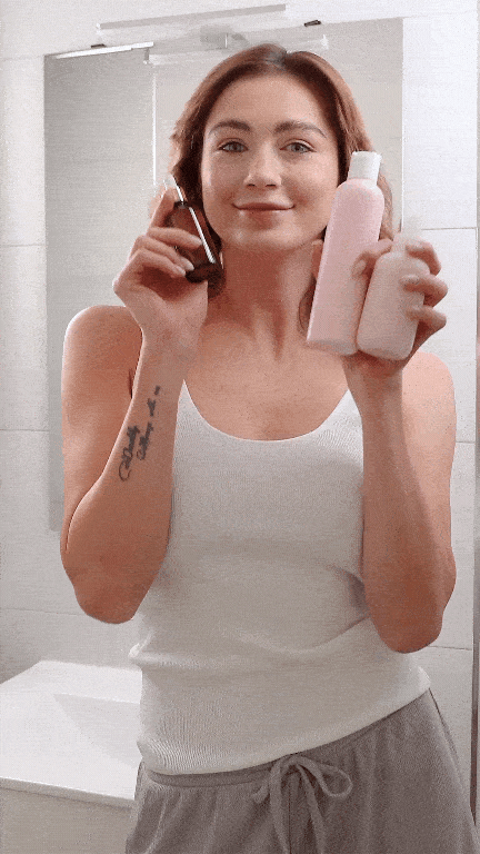 Young woman influencer smiles at the camera while holding three skincare bottles, in a bathroom setting. She is wearing a white tank top and gray pants, demonstrating the products in a casual, relatable manner.