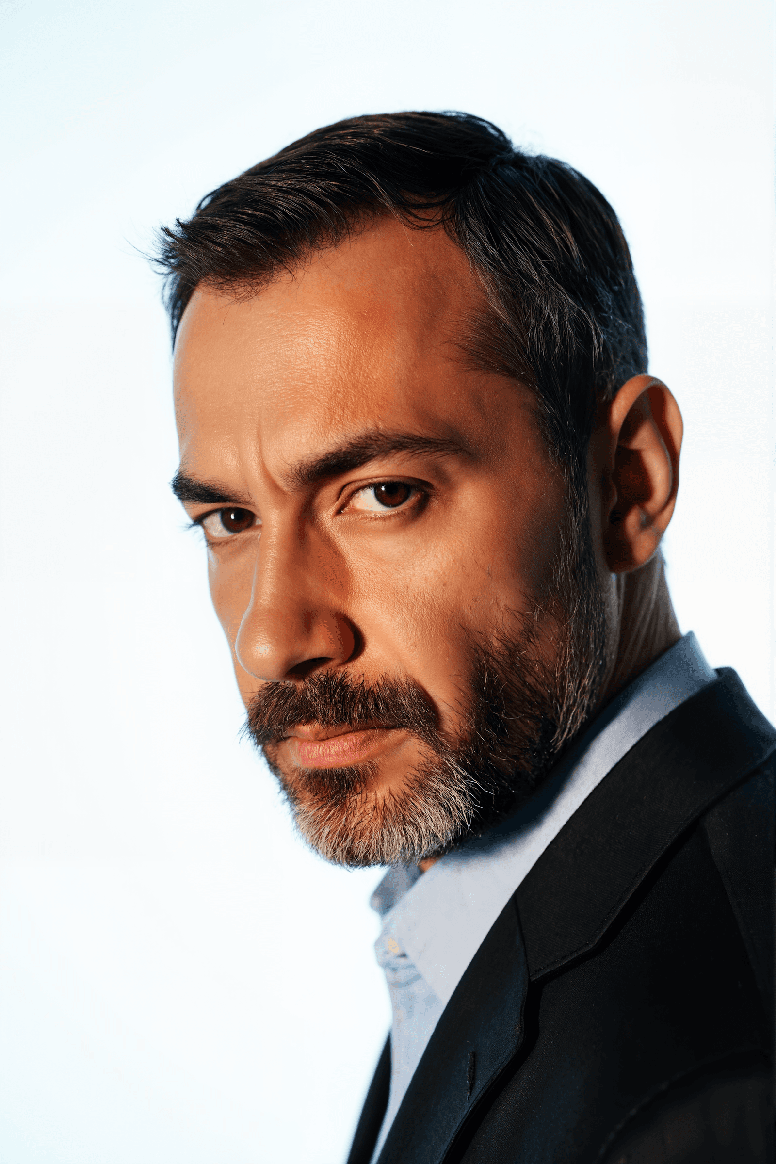Close-up of a man with a serious expression, brown eyes, and a neatly trimmed beard, wearing a suit jacket and light blue shirt.