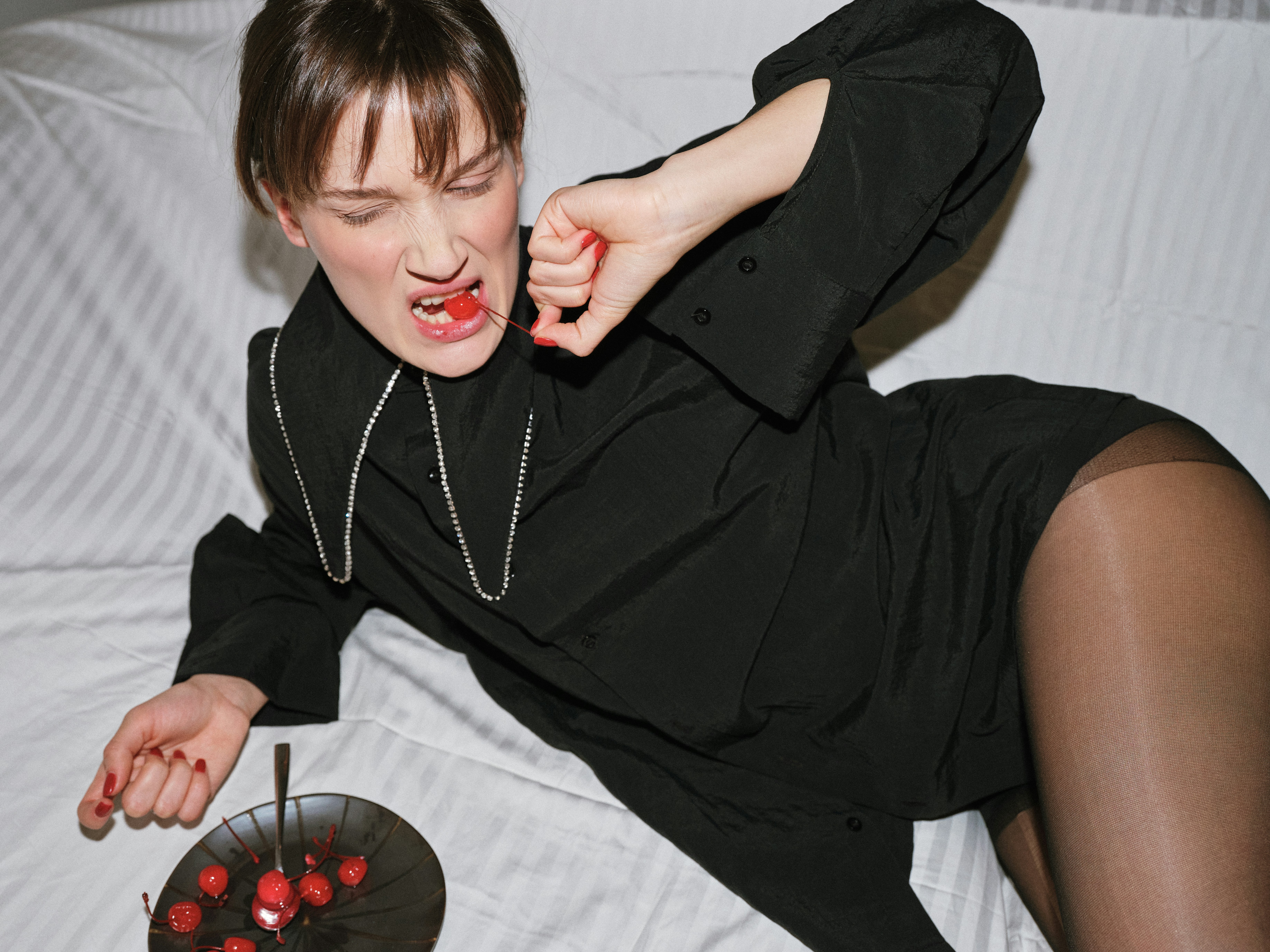 Dynamic editorial shot of a brunette model in a black shirt and sheer tights lying on a white bed, aggressively biting into a red cherry with a dark plate of cherries beside her