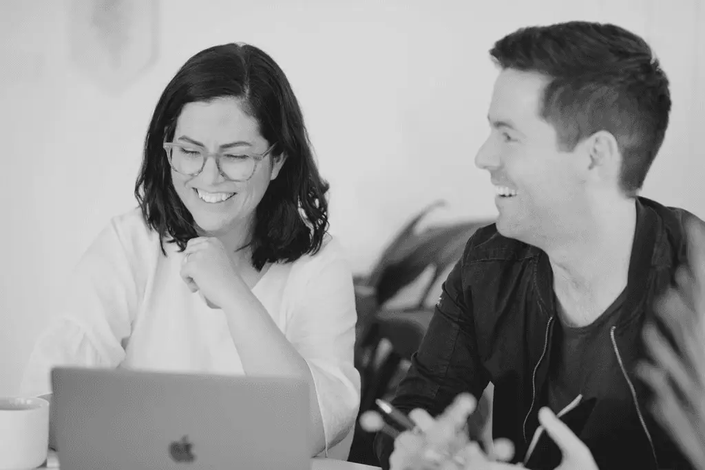 A black and white photo showing a smiling man and woman in conversation while working on a laptop.