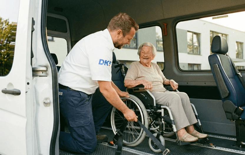 Krankenfahrer hilft älterer Frau im Rollstuhl aus dem Krankentransport