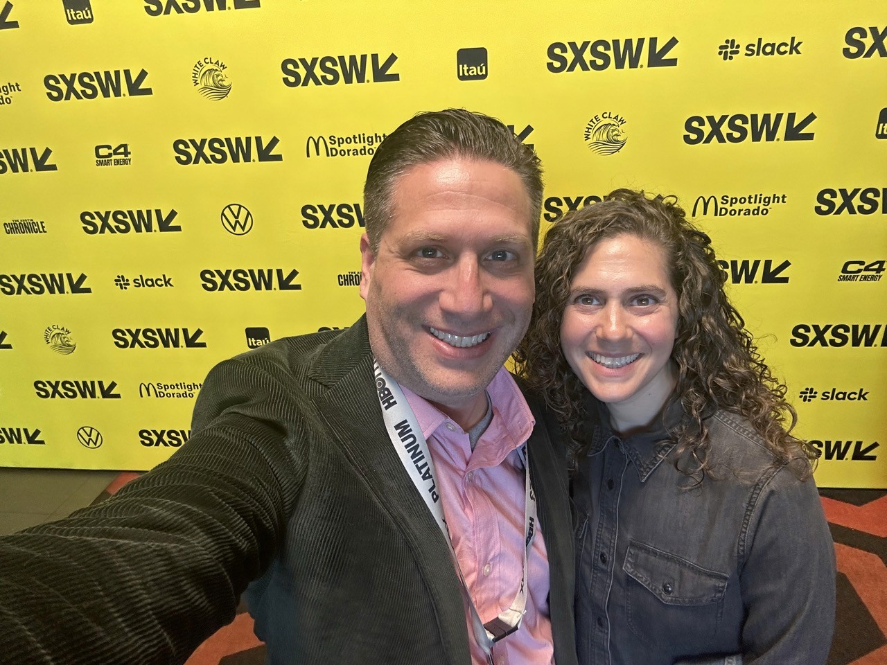 Shai and Becky Korman at SXSW, standing in front of a bright yellow backdrop covered with SXSW logos and sponsor names.