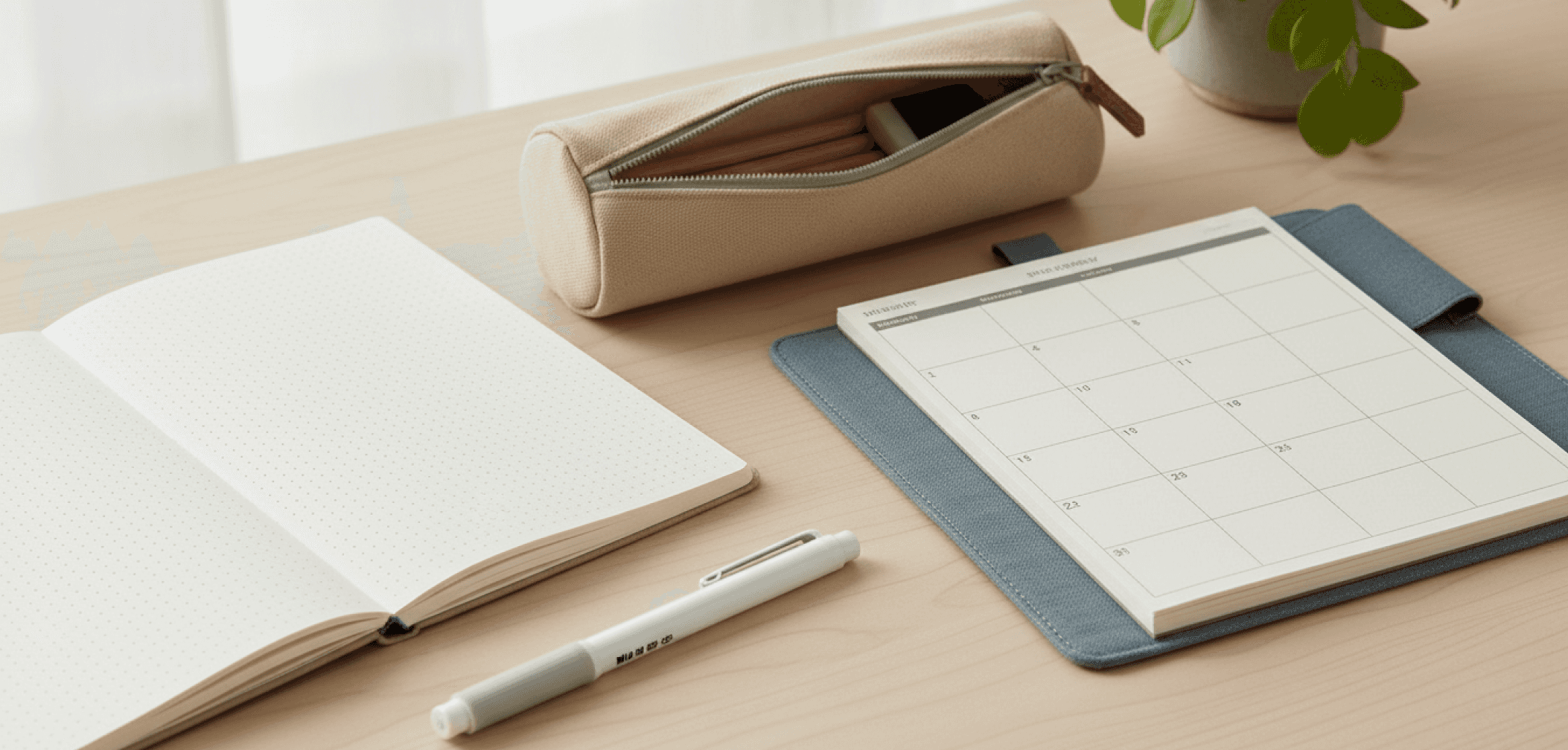 Minimal desk setup with an open notebook, calendar planner, white pen, beige pencil case, and small plant.