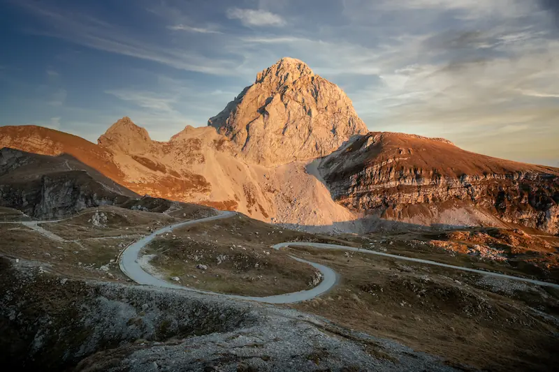 The serpentine mountain road at Mangart Saddle at sunset, leading toward the towering, sun-drenched peak of Mount Mangart in the Julian Alps.