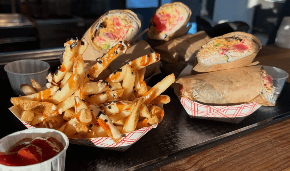 A rustic indoor scene featuring a table set for a meal, including two bowls of rice-based dishes topped with garnishes, an oil bottle, bread, a glass of juice, and milk bottles, all bathed in natural light streaming from a window.