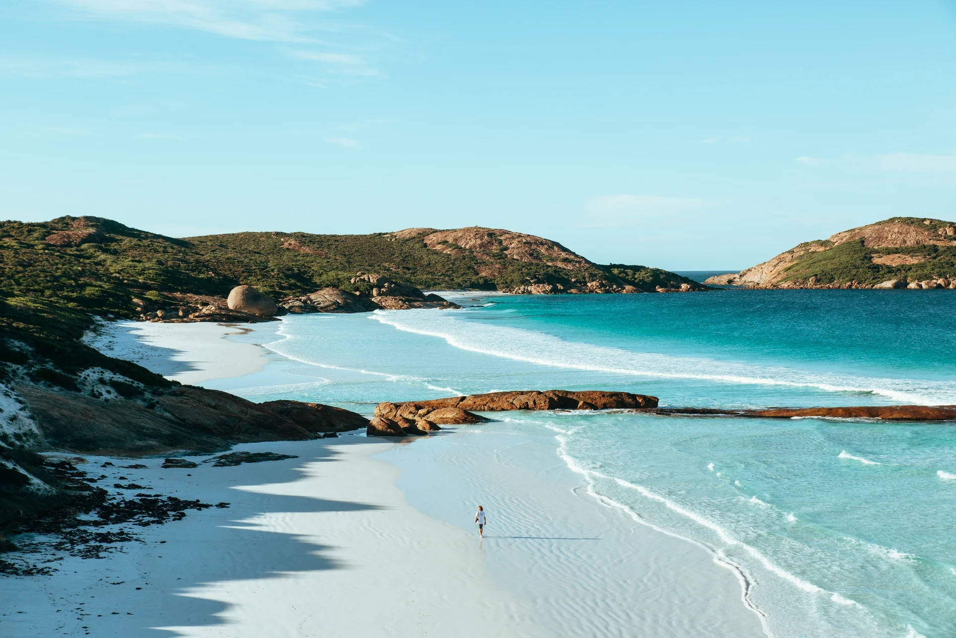 A person walking a wide white beach in a bay ringed by granite headlands