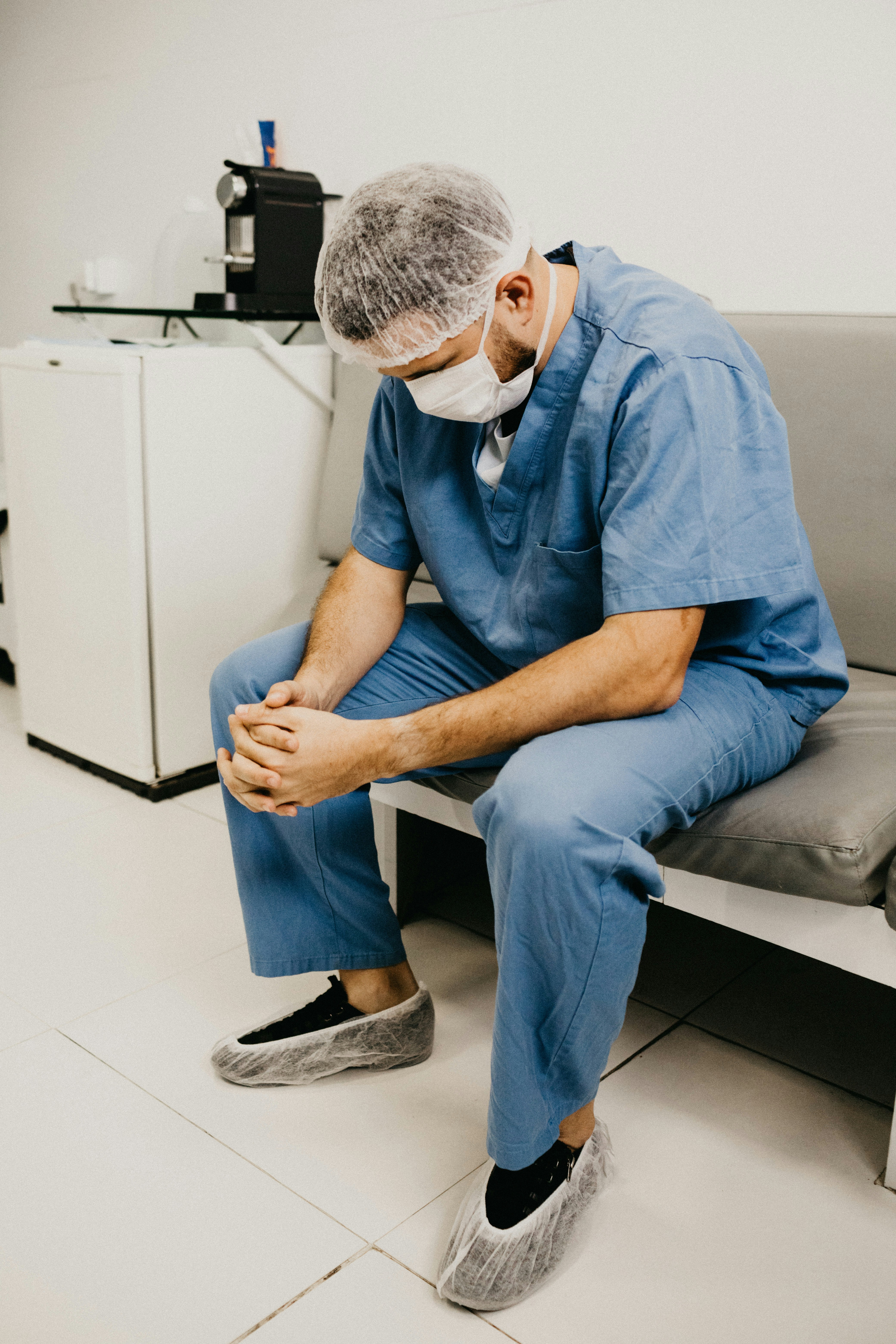 A healthcare professional in blue scrubs, a hair net, and a face mask sits on a bench with hands clasped, appearing thoughtful and reflective in a hospital break room.