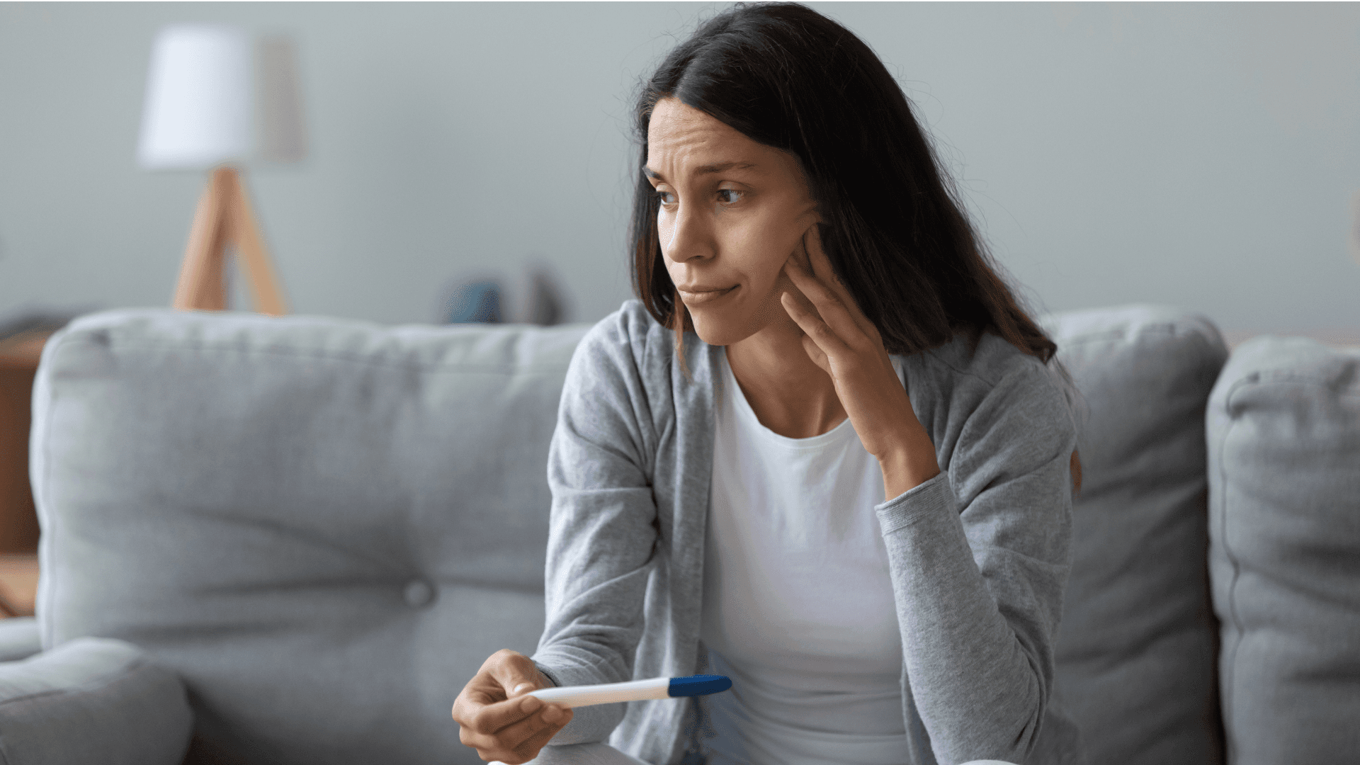 A woman sitting on a couch looks surprised or thoughtful while holding a pregnancy test.
