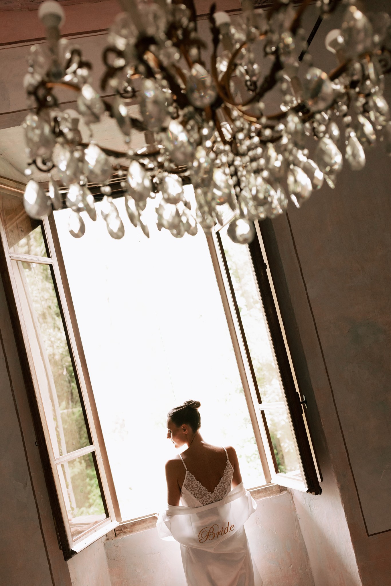 Eleonora in abito da sposa davanti alle finestre, lampadario di cristallo e luce naturale nei preparativi di matrimonio