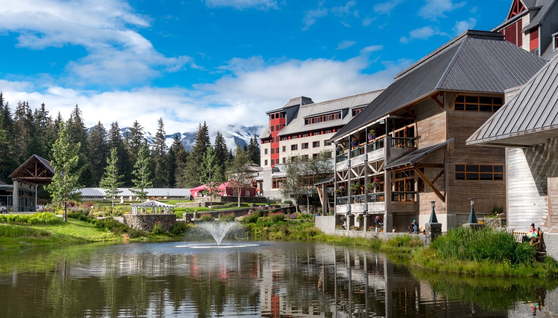 A multi-story lodge with wood and stone siding sits beside a pond with a fountain under a blue sky and pine trees.