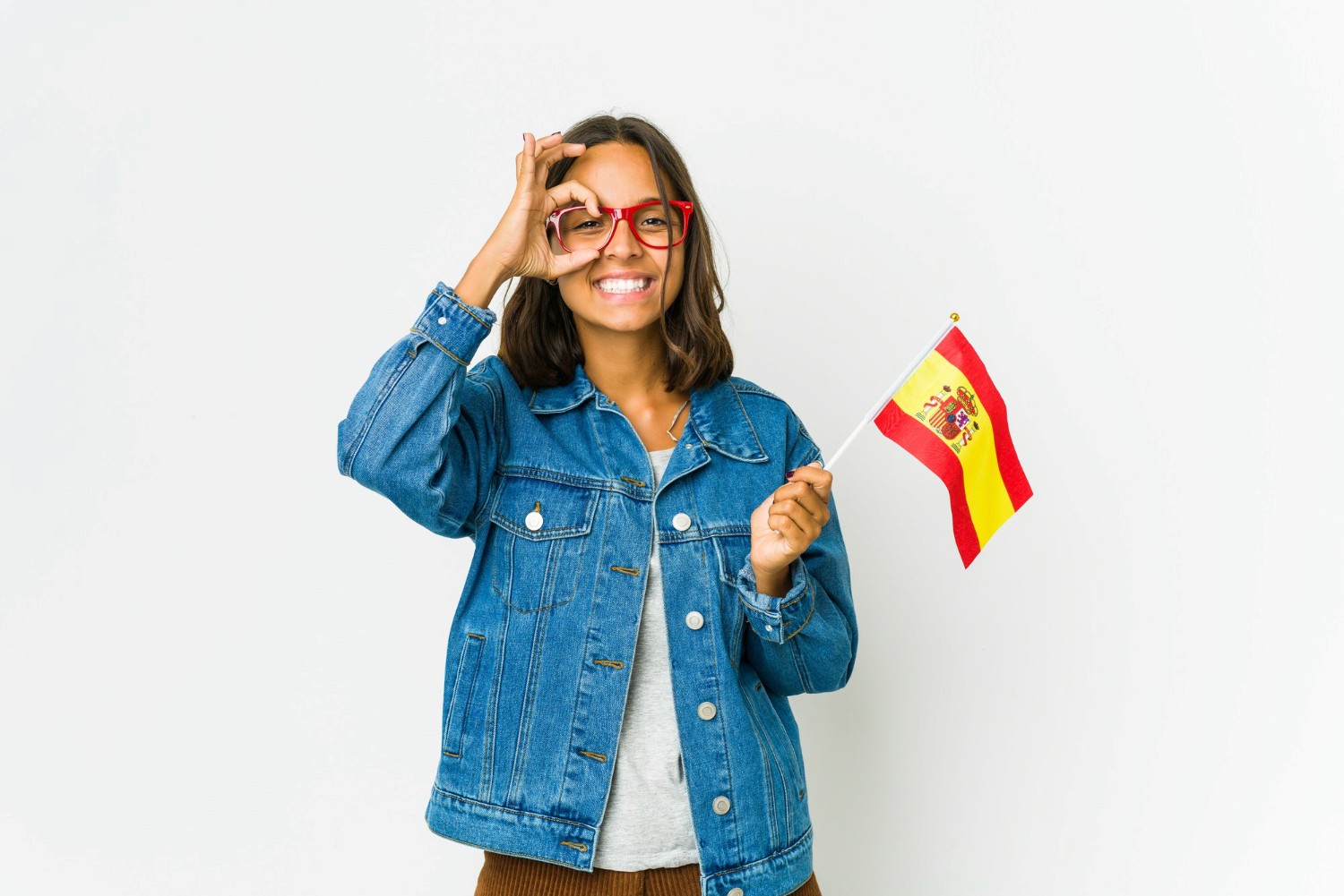 Mujer sonriente con gafas rojas y chaqueta de mezclilla sosteniendo una pequeña bandera de España, gesto de aprobación con la mano en un fondo blanco.
