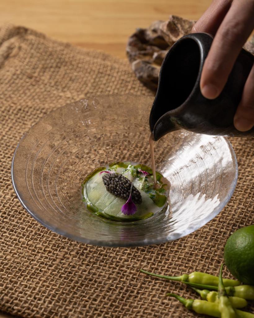 A hand pouring a sauce from a black vessel onto a plate containing caviar, herbs, and garnishes, surrounded by fresh ingredients on a textured burlap surface.