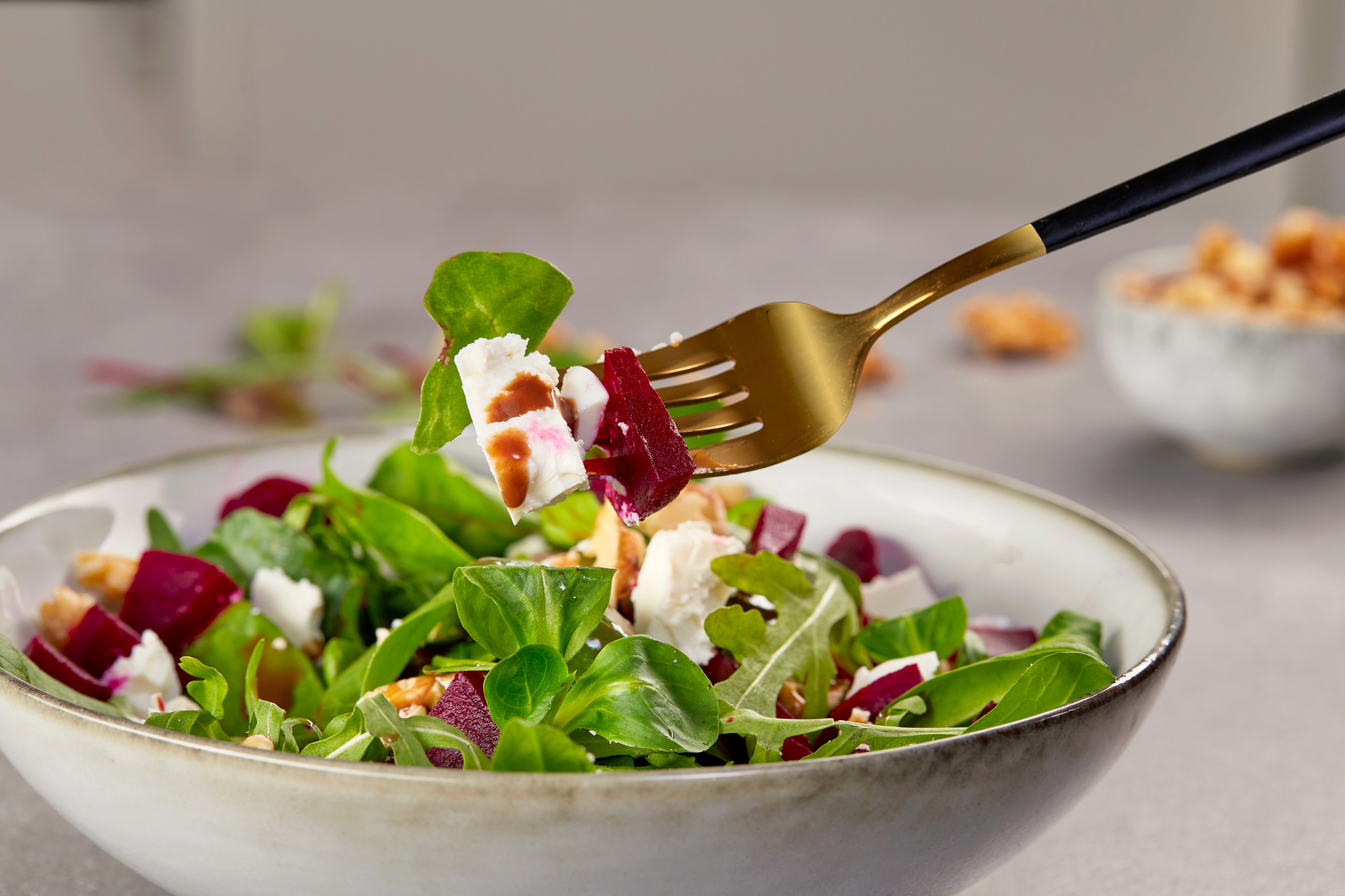 A fresh salad with vibrant beetroot, crumbled feta cheese, and mixed greens is lifted by a fork, showcasing a healthy and colorful dish in a ceramic bowl on a textured surface.