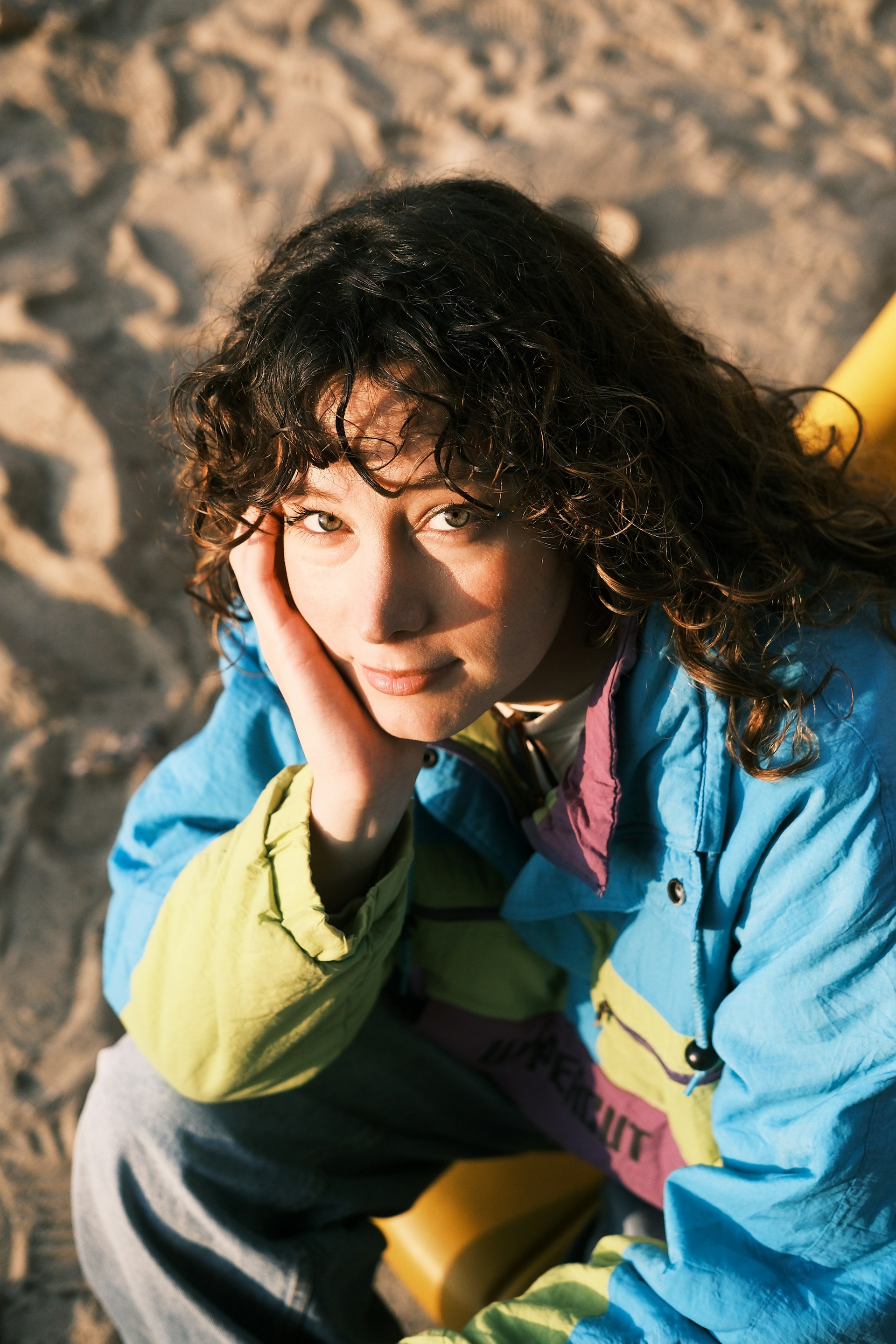 A woman with curly hair wearing a bright, multi-colored vintage windbreaker sitting on a beach. She is resting her chin on her hand and looking directly at the camera.