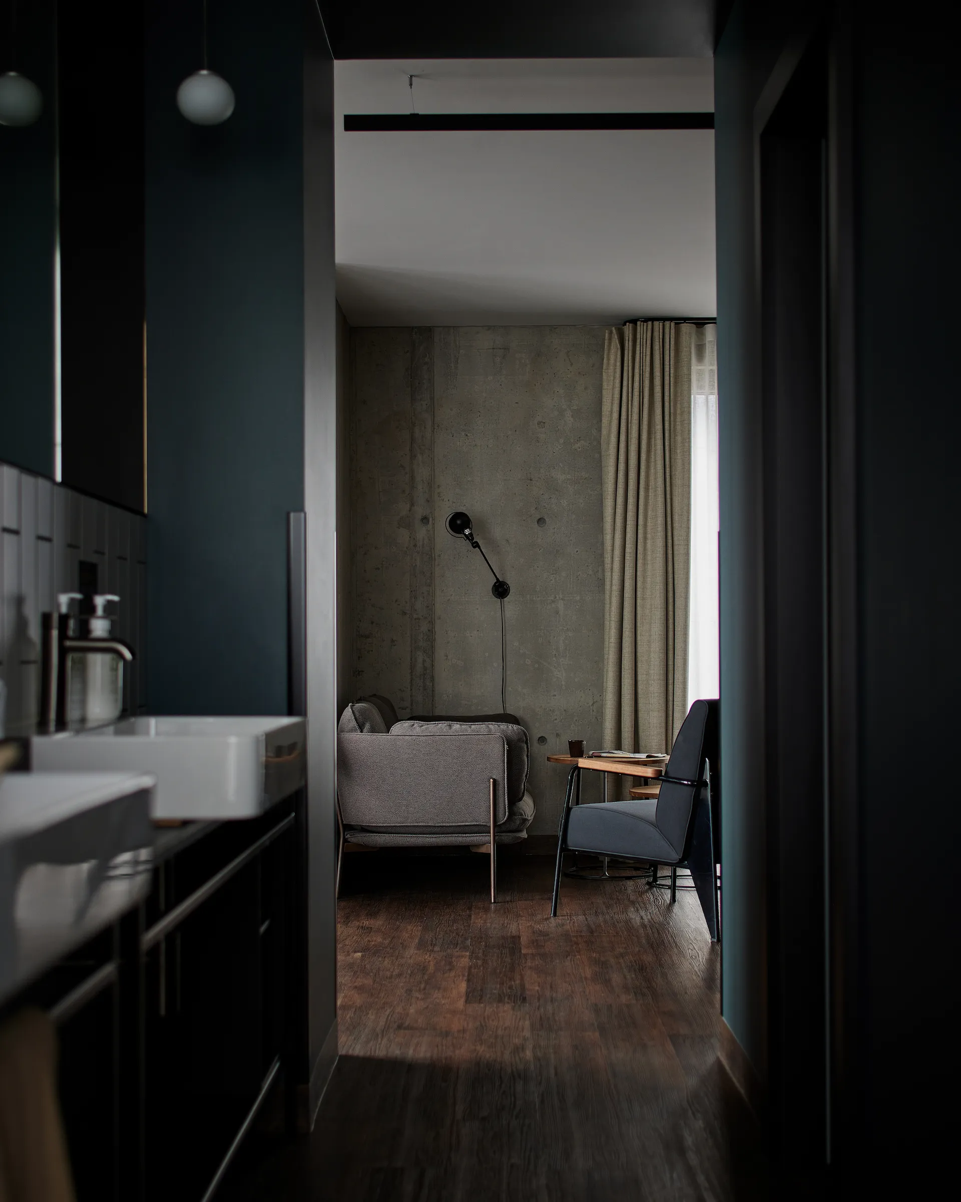 View from the Harbour Suite’s dark green bathroom through to the living area, with a grey sofa, black chair and linen curtain at the window