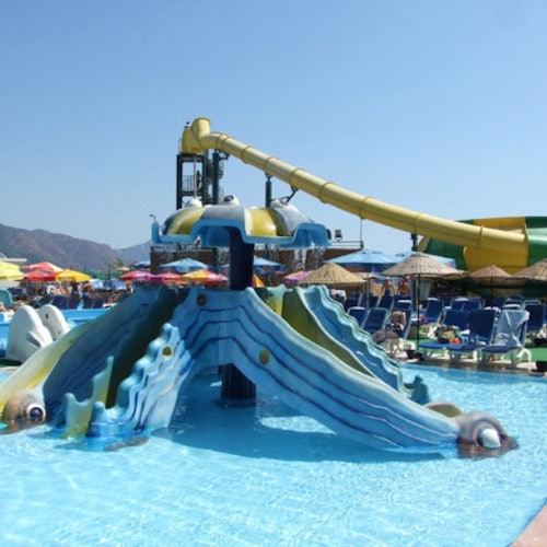 Colorful water park with fish-themed slides, a large yellow slide in the background, and umbrellas and lounge chairs around.