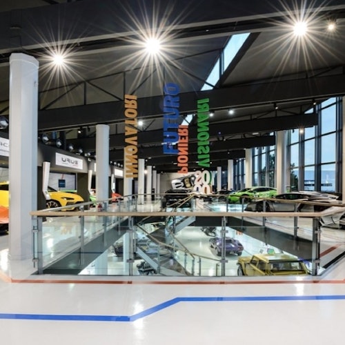 Sala de exhibición moderna con elegantes autos deportivos en exhibición, luces de techo iluminadas, barandillas de vidrio y letreros colgantes coloridos.