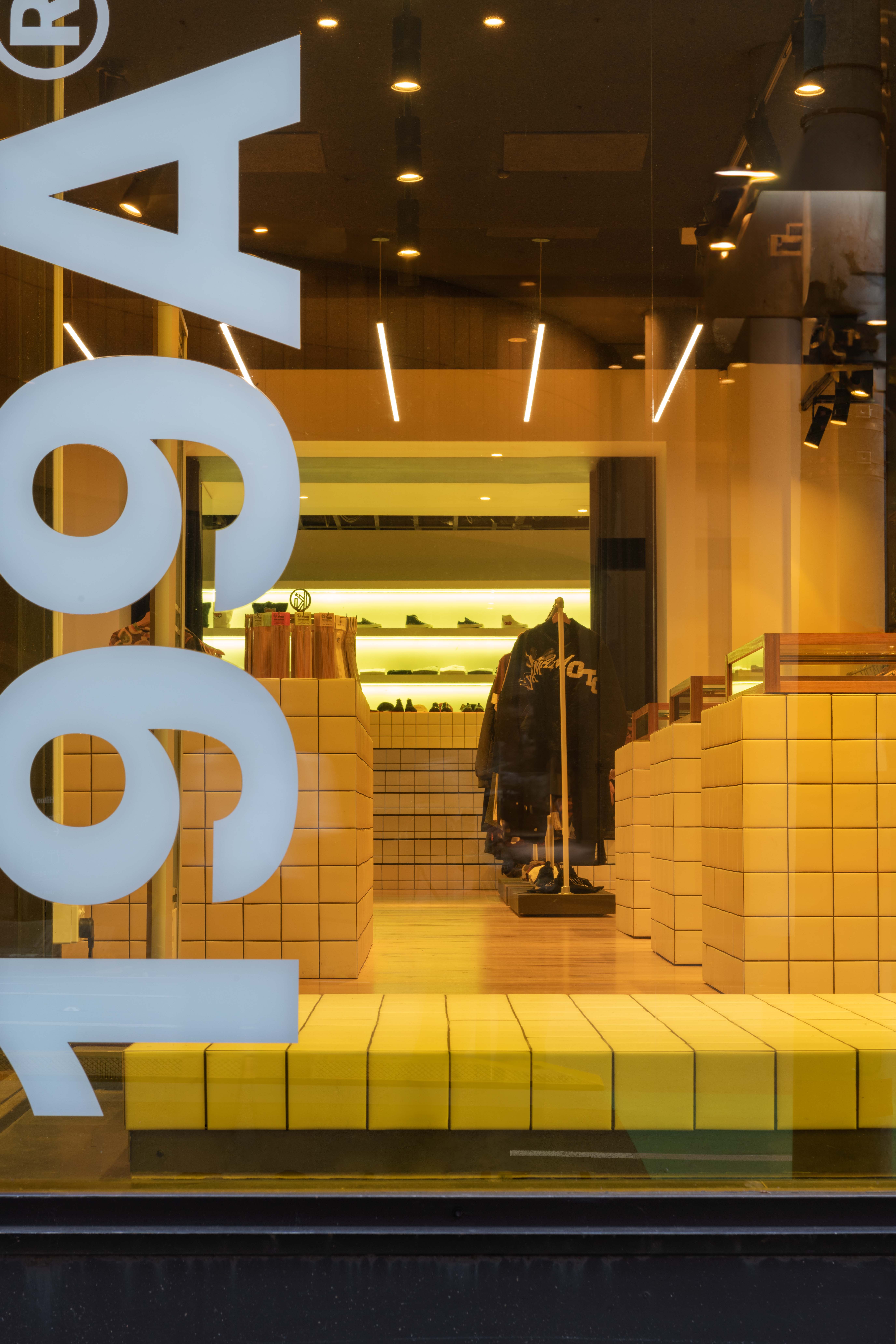 Street-level view through the 199A® storefront window, revealing a warm-lit interior with tiled plinths, stacked display blocks, hanging garments, and graphic signage layered across the glass.