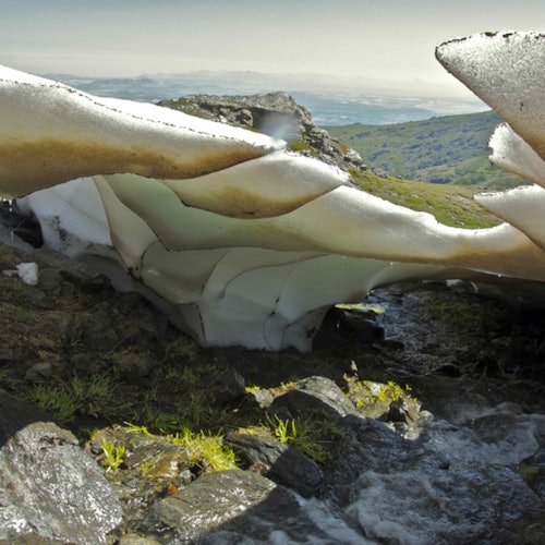 在晴朗的天空下，大片的冰覆盖着岩石溪流，间或点缀着草地，背景是一片丘陵地貌。