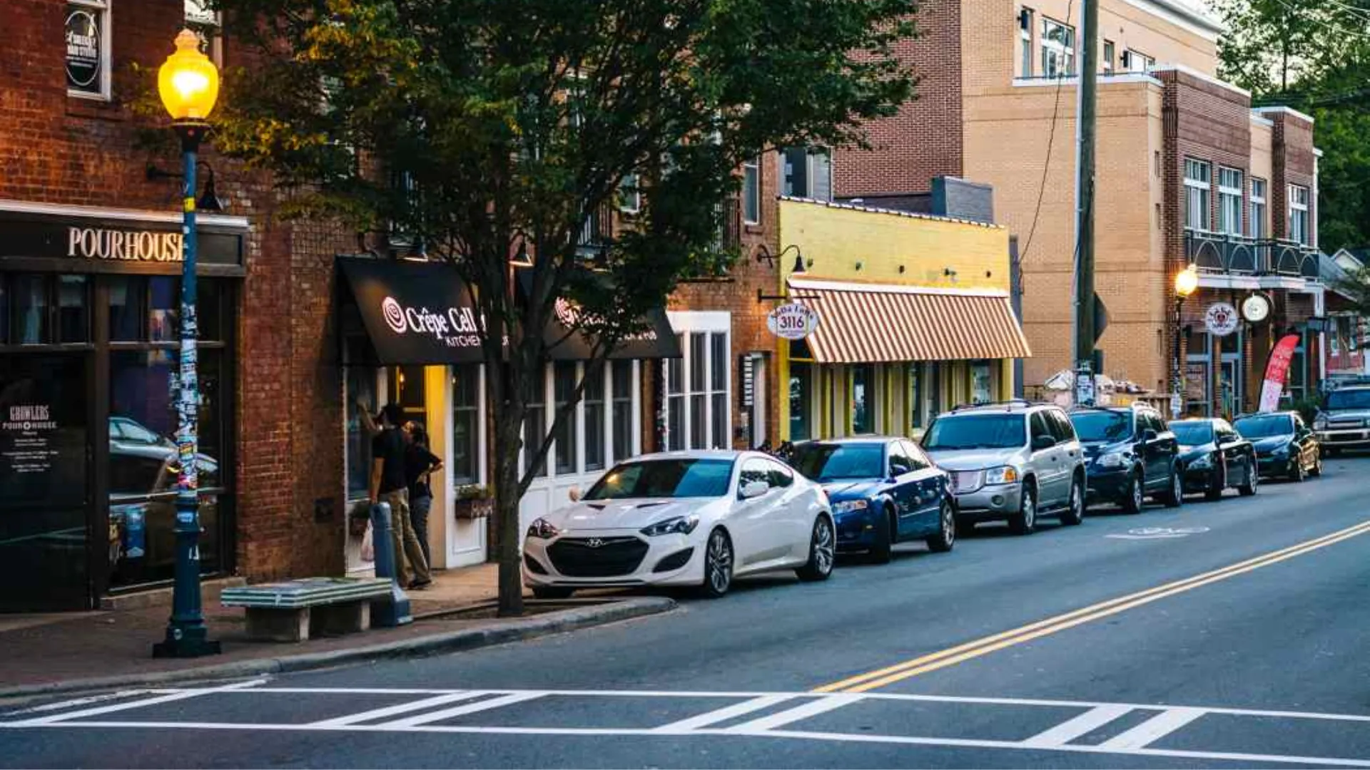 A busy street scene featuring parked cars and pedestrians walking along the sidewalk.