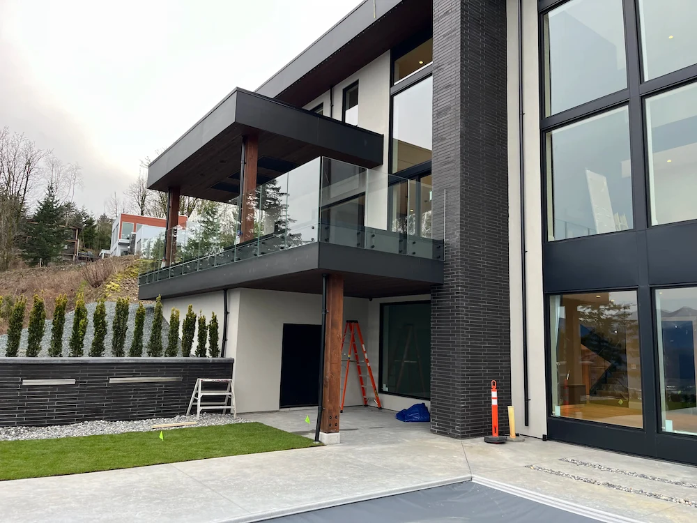 Modern two-storey home with frameless glass balcony railing and exposed timber posts, installed on a sloped property in British Columbia.