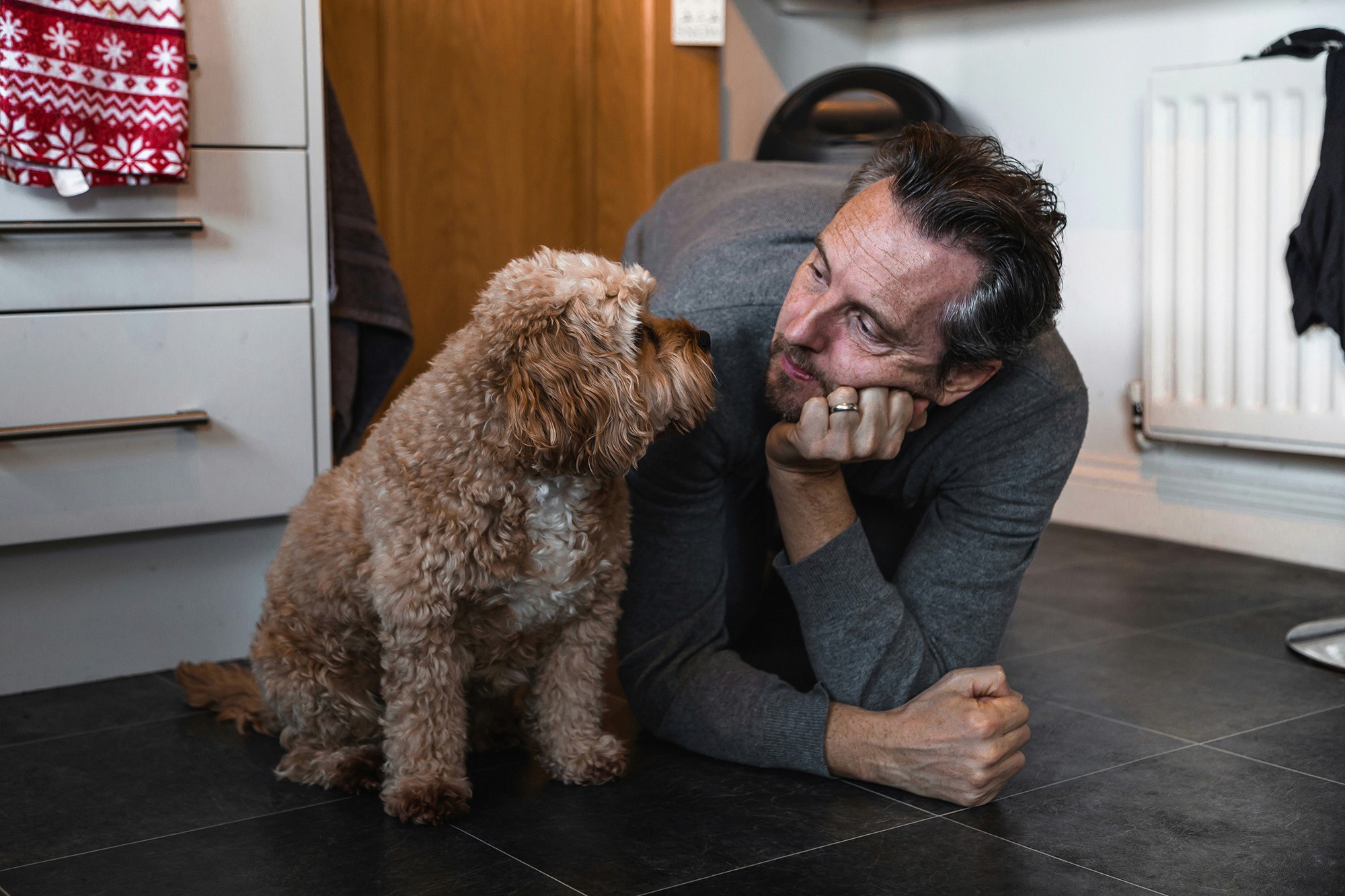 Man lying on the floor, smiling and making eye contact with his curly-haired dog in a cozy kitchen setting