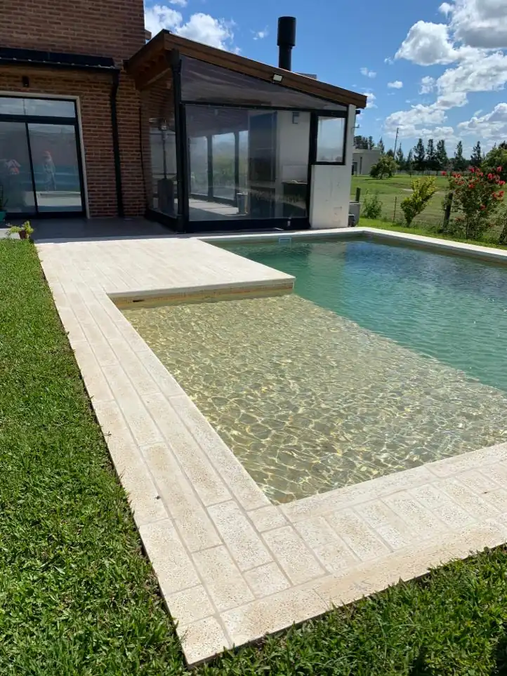 Pileta de hormigón con un extenso solárium húmedo revestido en travertino, ubicada estratégicamente junto a una casa con quincho vidriado.