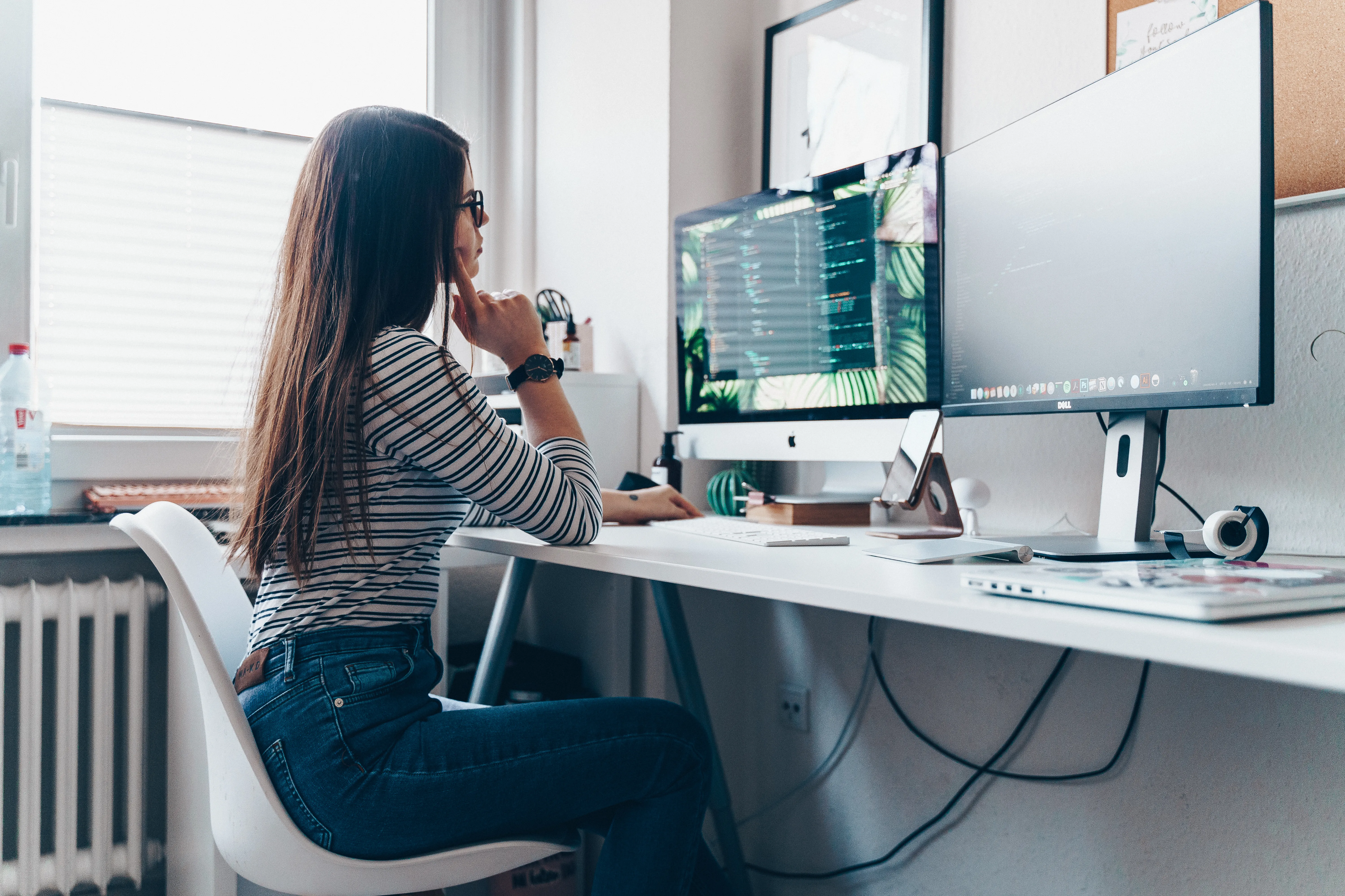 Woman sitting at a desk with dual monitors, focused on writing code in a modern home office setup