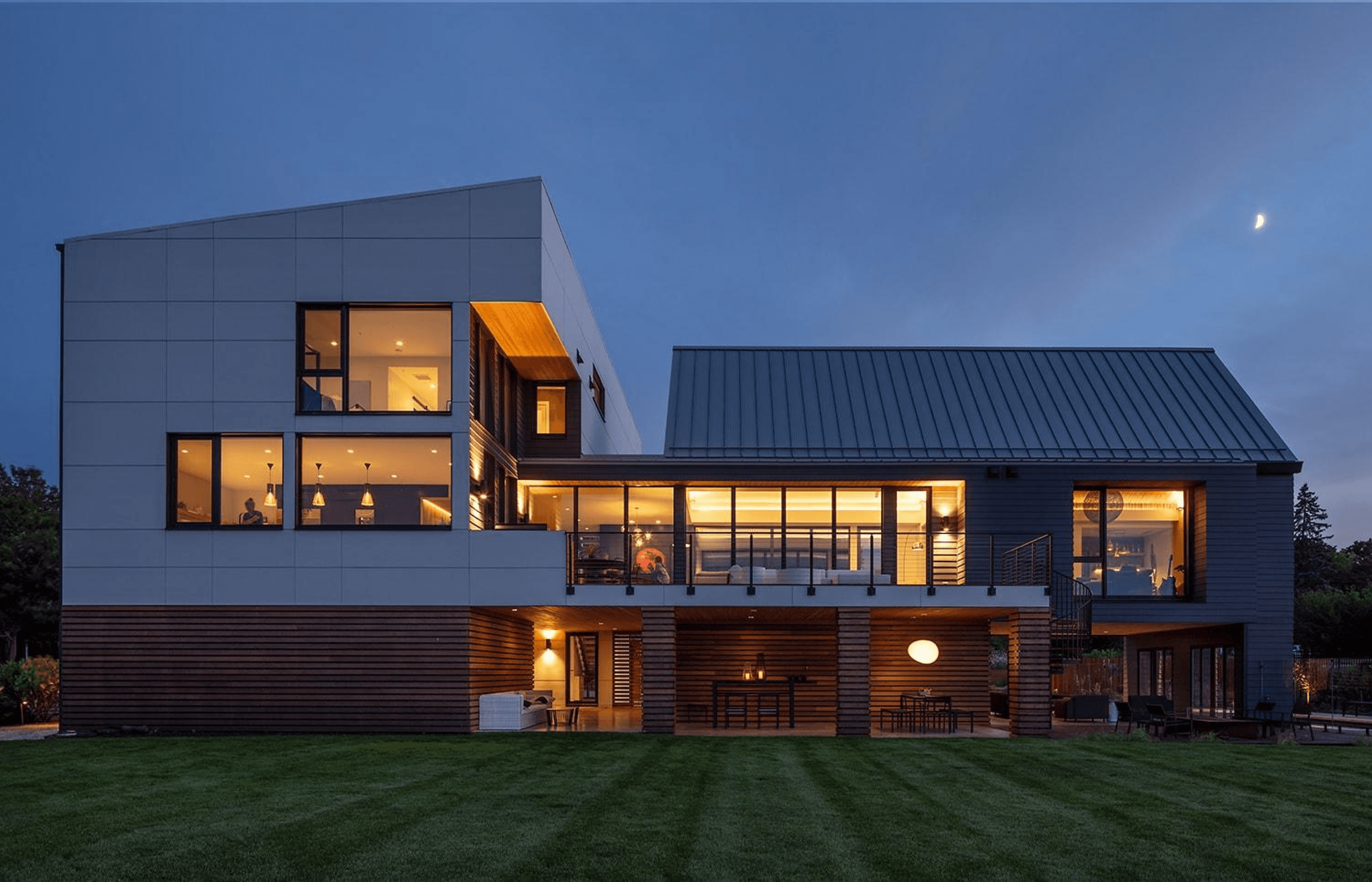 Modern two-story house at dusk, featuring large windows and a well-lit interior, surrounded by a grassy lawn.