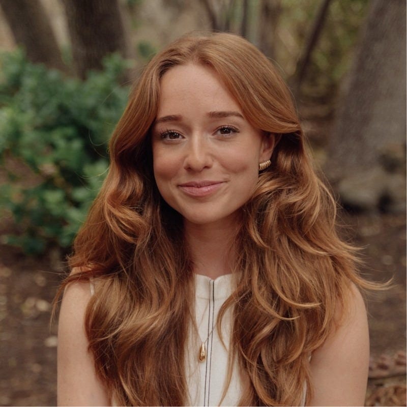 Smiling woman in beige linen outfit standing indoors with plants and books in the background.