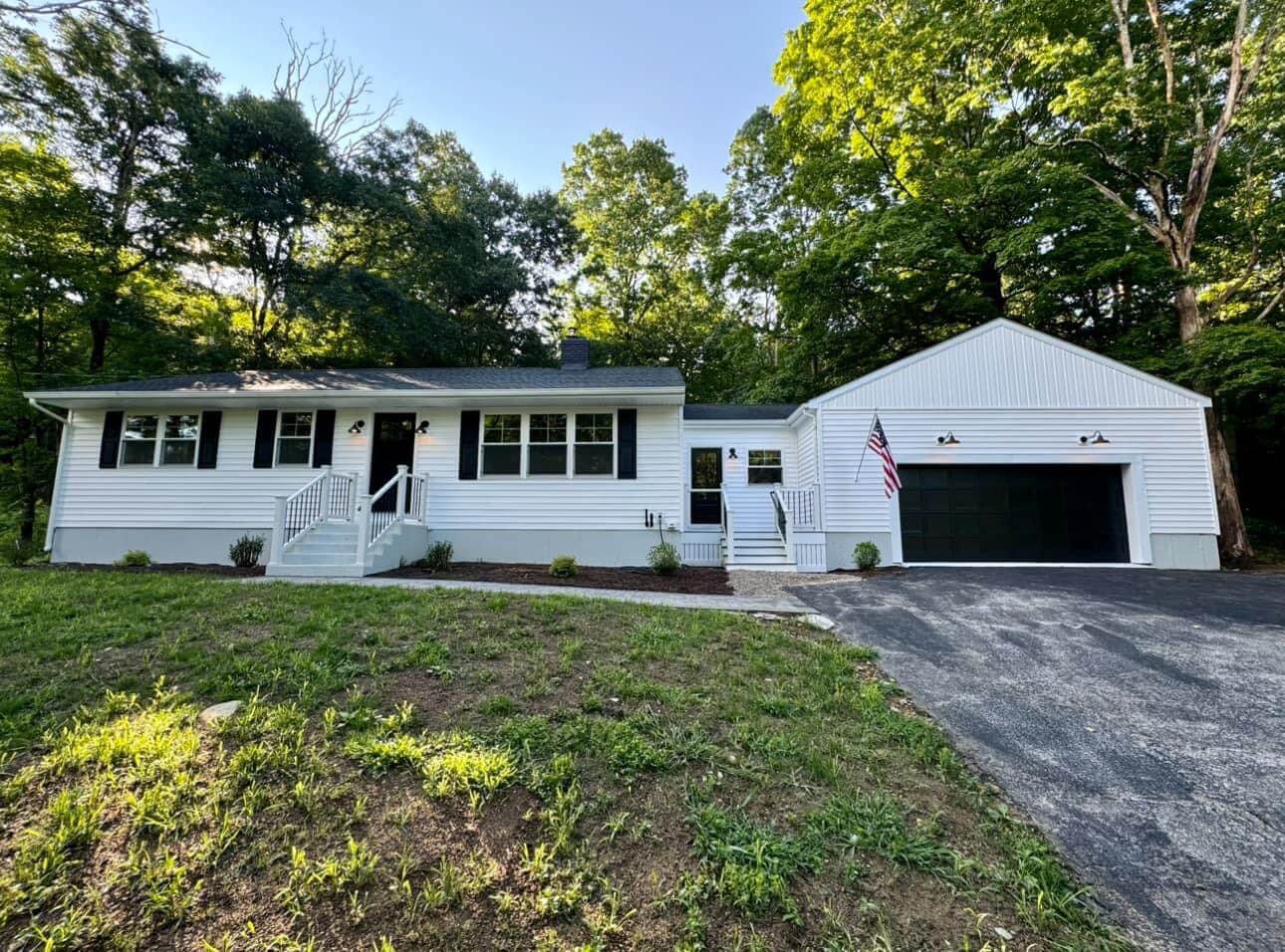 A single-story white home with black shutters and a matching black garage door, framed by a lush wooded background. The house features a simple front porch with steps leading to the entrance and a paved driveway.