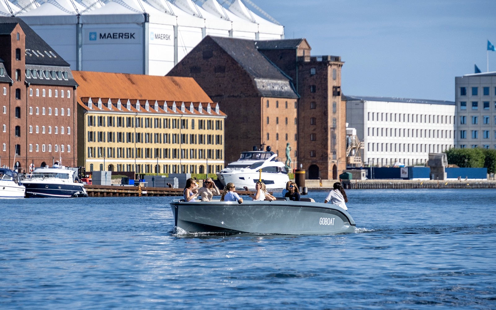 Vendégek egy hajókiránduláson Koppenhága kikötőjében, ahol a háttérben történelmi épületek láthatók.
