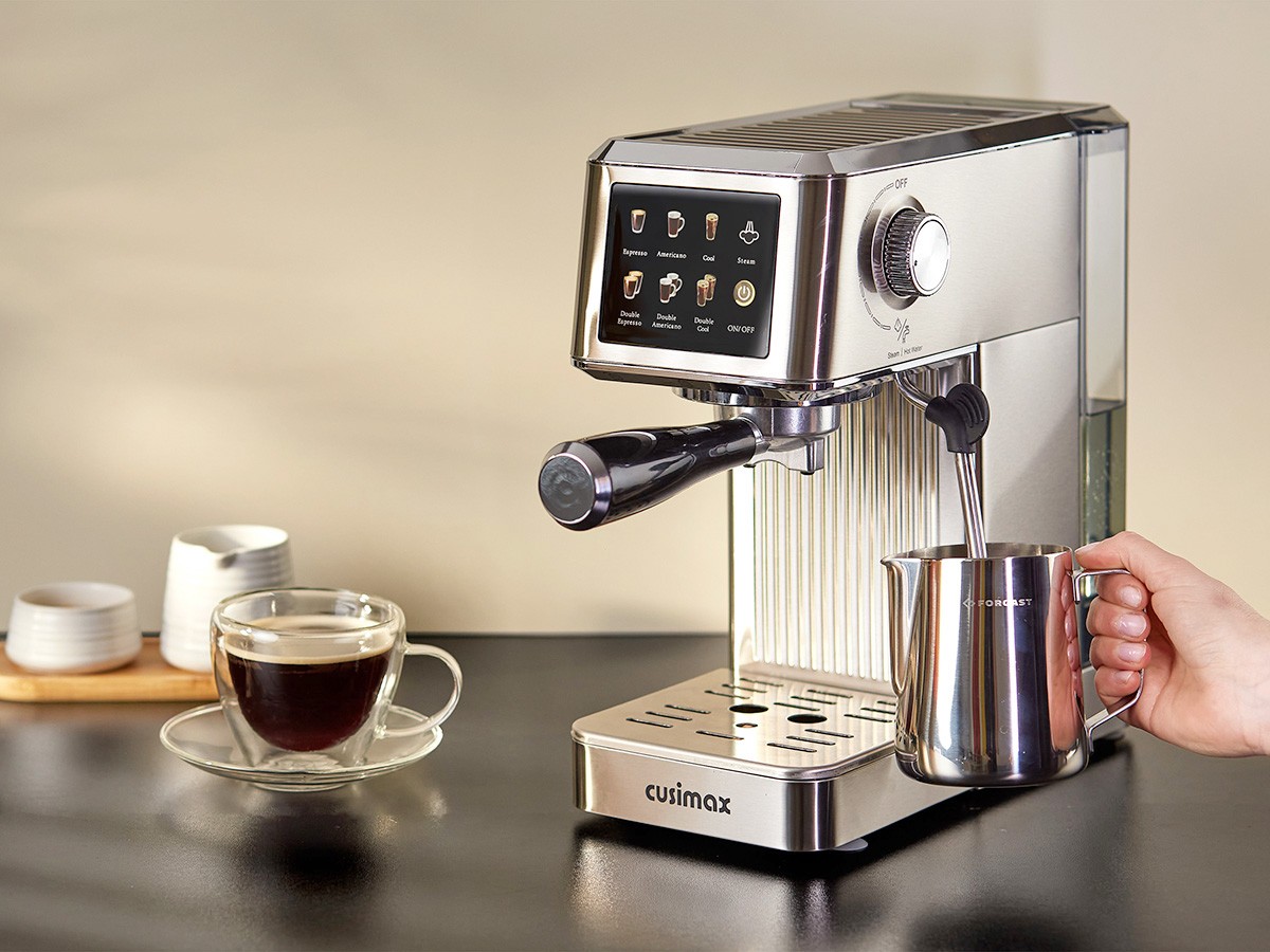 A modern stainless steel espresso machine with a digital display is steaming milk into a metal frothing pitcher, while a glass cup of black coffee sits on a nearby countertop, next to a small stack of saucers and bowls, bathed in warm light.