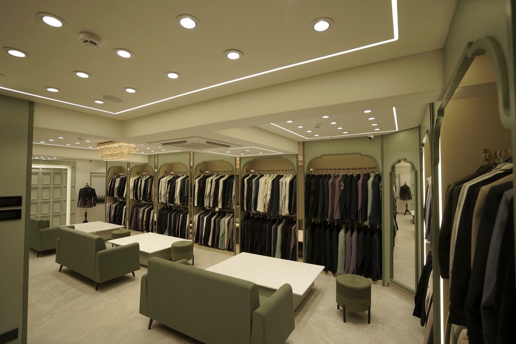 Jalaram Wedding Couture: seating area with sage-green sofas and white tables in front of garment racks, by The Auura